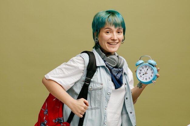 Sonriente joven estudiante con auriculares y pañuelo en el cuello y mochila de pie en la vista de perfil agarrando mochila mirando a la cámara que muestra el despertador aislado en el fondo verde oliva