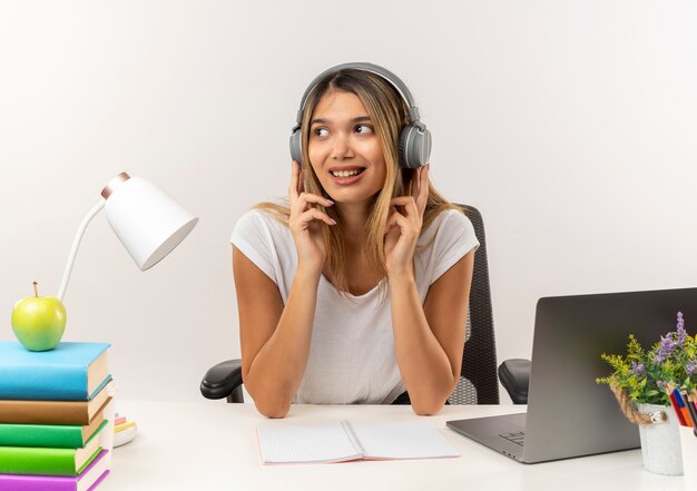 Sonriente joven estudiante bonita con audífonos sentado en el escritorio con herramientas escolares escuchando música mirando al lado con los dedos en los auriculares aislados en la pared blanca