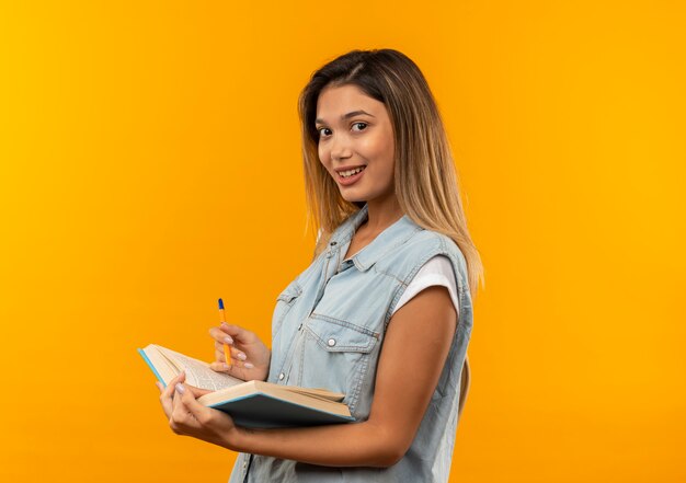 Sonriente joven estudiante bonita vestida con bolsa trasera de pie en la vista de perfil sosteniendo el libro abierto y la pluma aislada en la pared naranja