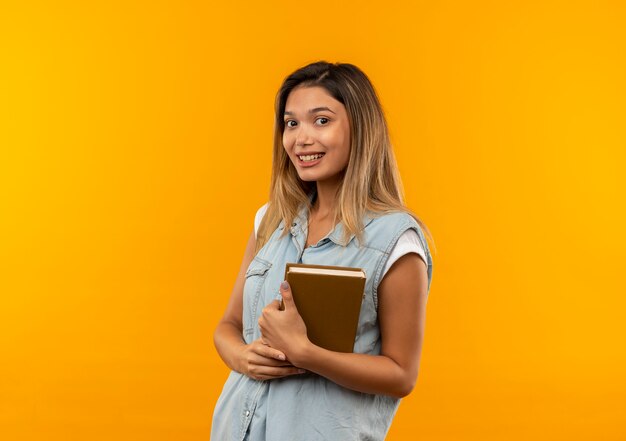 Sonriente joven estudiante bonita vestida con bolsa trasera sosteniendo el libro aislado en la pared naranja