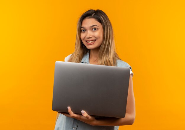 Sonriente joven estudiante bonita vestida con bolsa trasera sosteniendo portátil aislado en la pared naranja
