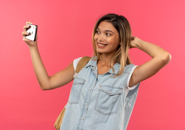 Foto gratuita sonriente joven estudiante bonita vistiendo bolsa trasera poniendo la mano detrás de la cabeza y tomando selfie aislado en la pared rosa