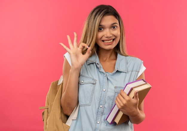 Sonriente joven estudiante bonita vistiendo bolsa trasera sosteniendo libros y haciendo bien signo aislado en la pared rosa