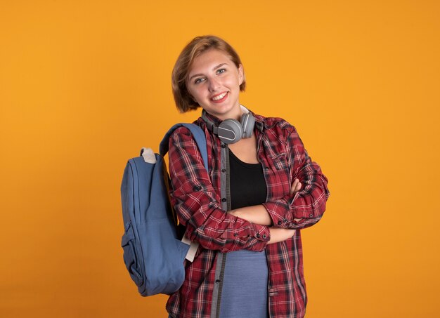 Sonriente joven estudiante eslava con auriculares con mochila se encuentra con los brazos cruzados.