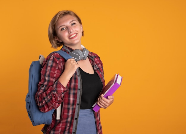 Sonriente joven estudiante eslava con auriculares con mochila se encuentra lateralmente sosteniendo el libro y el cuaderno