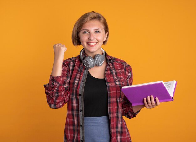 Sonriente joven estudiante eslava con auriculares con mochila mantiene el puño sostiene el libro