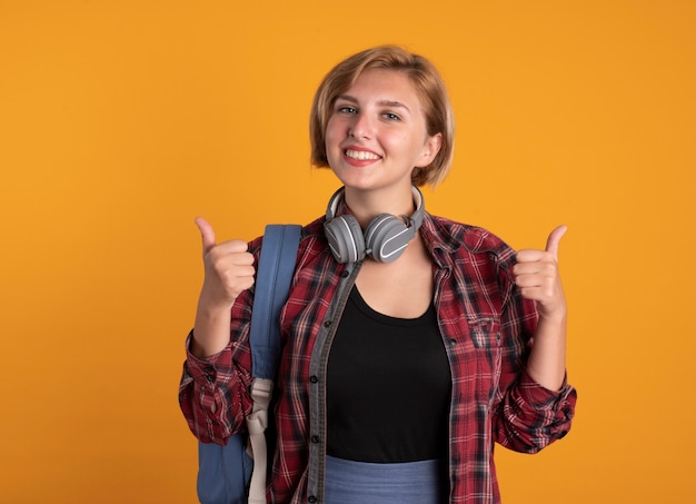 Sonriente joven estudiante eslava con auriculares con mochila pulgar hacia arriba de las dos manos