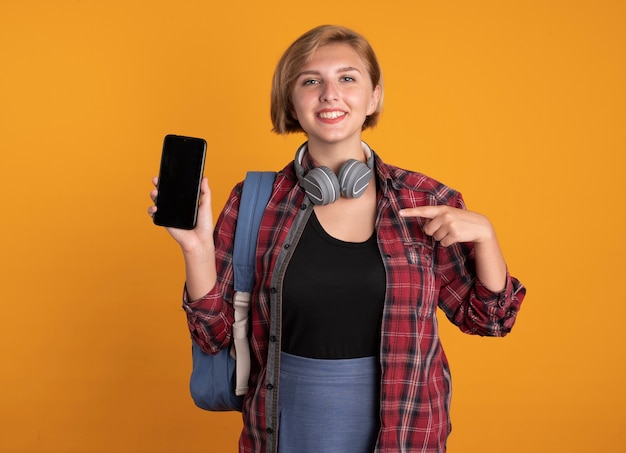 Sonriente joven estudiante eslava con auriculares con mochila sostiene y apunta al teléfono