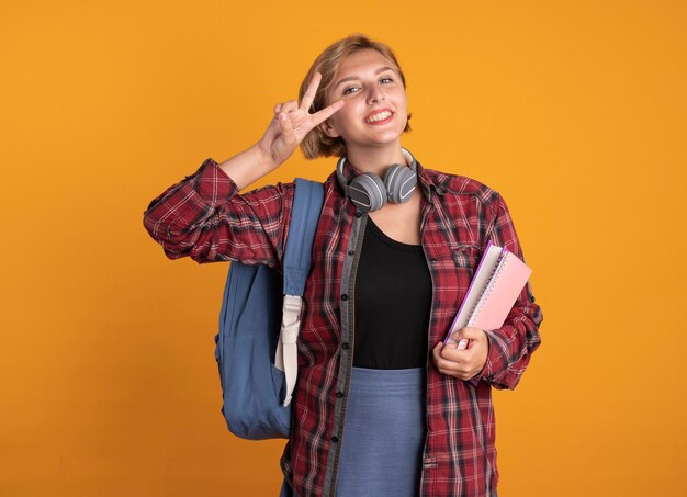 Sonriente joven estudiante eslava con auriculares con mochila sostiene gestos de libro y portátil signo de mano de victoria