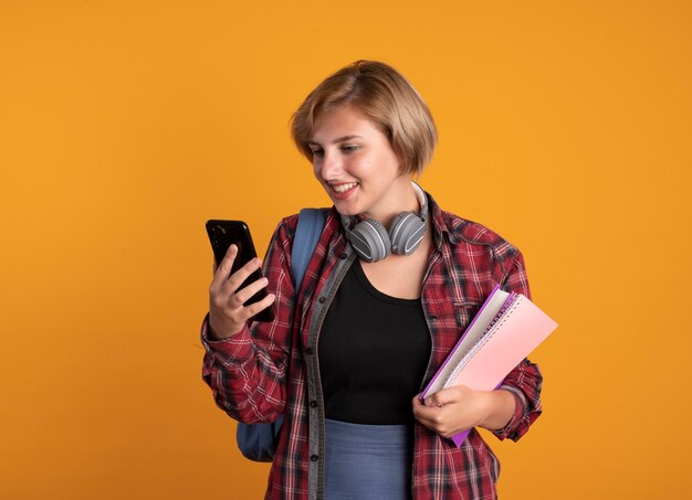 Sonriente joven estudiante eslava con auriculares con mochila sostiene libro y portátil mira teléfono