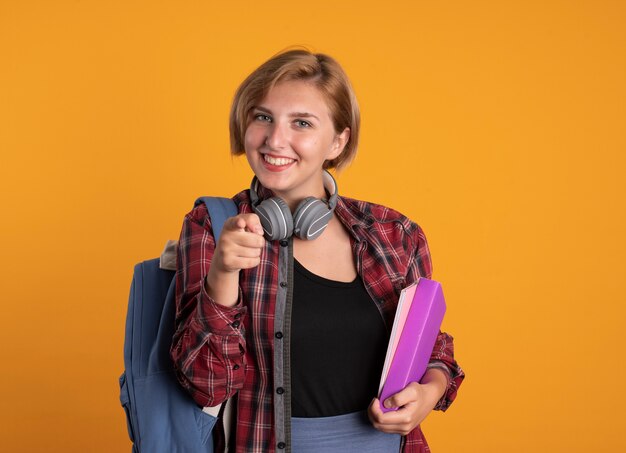 Sonriente joven estudiante eslava con auriculares con mochila sostiene puntos de libro y cuaderno a la cámara