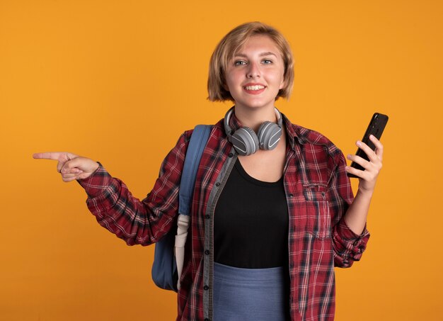 Sonriente joven estudiante eslava con auriculares con mochila sostiene el teléfono y apunta al lado