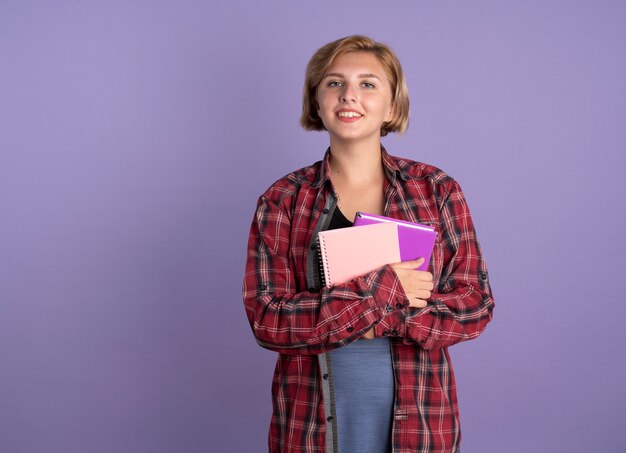 Sonriente joven estudiante eslava sostiene libro y cuaderno