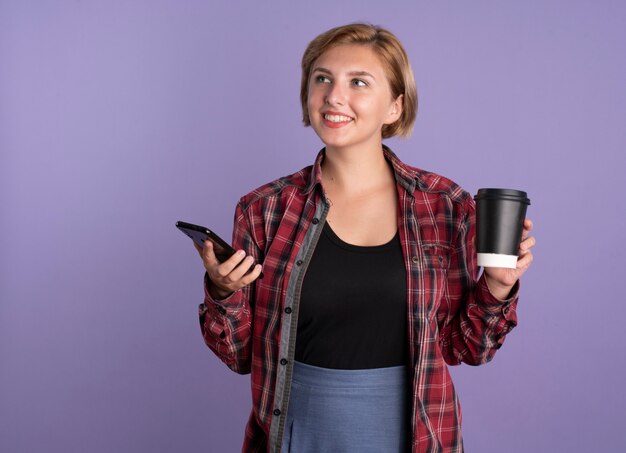 Sonriente joven estudiante eslava sostiene el teléfono y la taza mirando al lado