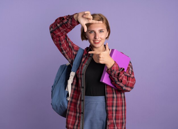 Sonriente joven estudiante eslava vestida con mochila gestos marco con las manos sosteniendo el libro y el cuaderno