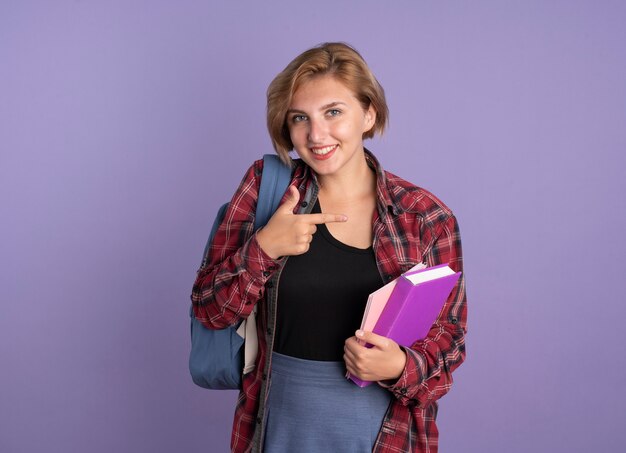 Sonriente joven estudiante eslava vestida con mochila sosteniendo el libro y el cuaderno apuntando al lado