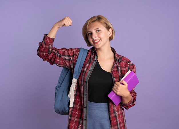 Sonriente joven estudiante eslava vestida con mochila tensa bíceps sostiene libro y cuaderno