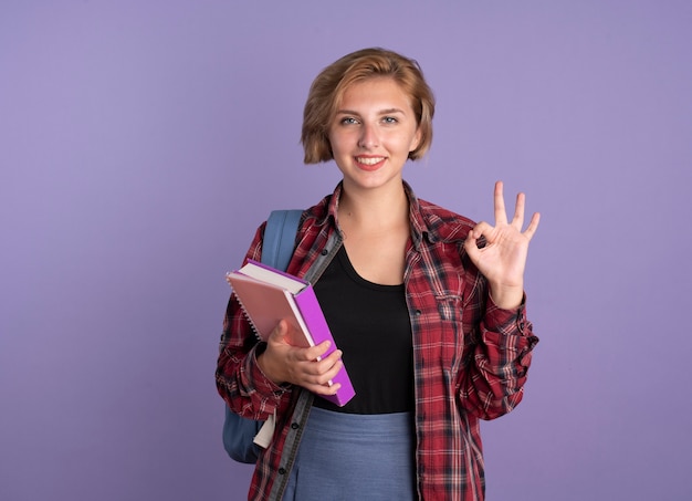 Sonriente joven estudiante eslava vestida con mochila tiene gestos de libro y cuaderno bien signo de mano