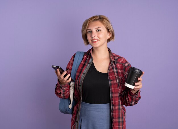 Sonriente joven estudiante eslava vestida con mochila tiene taza de papel y teléfono