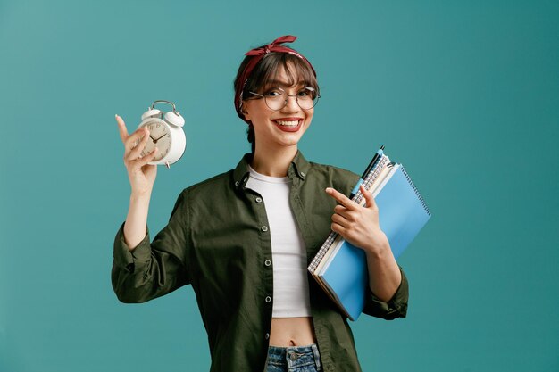 Sonriente joven estudiante con gafas de bandana sosteniendo grandes blocs de notas con bolígrafo mirando a la cámara mostrando el despertador apuntando al reloj aislado en el fondo azul