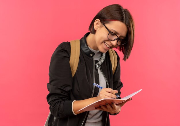 Sonriente joven estudiante con gafas y bolsa trasera escribiendo algo en el bloc de notas aislado en la pared rosa