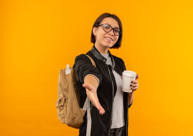 Sonriente joven estudiante con gafas y bolsa trasera sosteniendo una taza de café de plástico extendiendo la mano hacia el frente gesticulando Hola aislado en la pared naranja