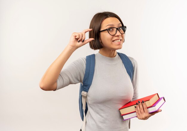 Sonriente joven estudiante con gafas y mochila sosteniendo libros que muestran el tamaño aislado en la pared blanca