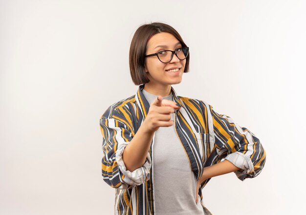 Sonriente joven estudiante con gafas poniendo la mano en la cintura y apuntando al frente aislado en la pared blanca