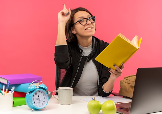 Sonriente joven estudiante con gafas sentado en el escritorio con herramientas universitarias sosteniendo el libro haciendo la tarea con el dedo levantado aislado en la pared rosa