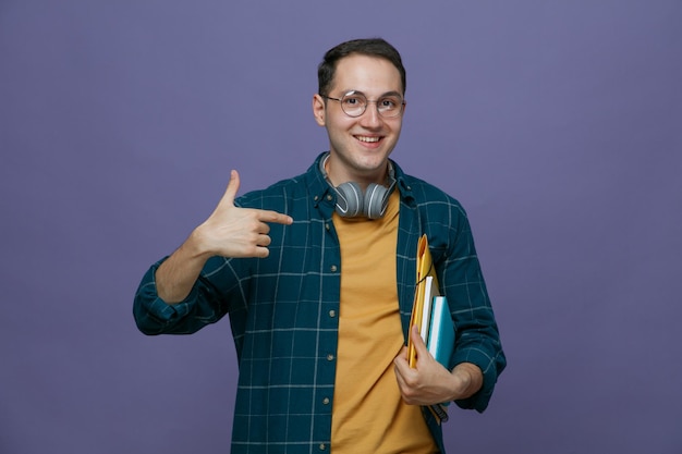 Sonriente joven estudiante masculino con anteojos auriculares alrededor del cuello sosteniendo una carpeta de notas bajo el brazo apuntándolos mirando la cámara aislada en un fondo morado