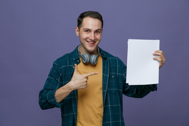 Foto gratuita sonriente joven estudiante masculino usando audífonos alrededor del cuello mirando a la cámara mostrando los exámenes apuntándolos aislados en un fondo morado