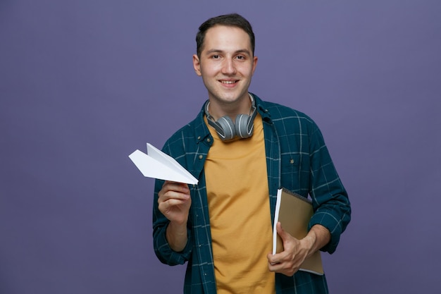 Sonriente joven estudiante masculino usando audífonos alrededor del cuello sosteniendo un bloc de notas bajo el brazo y un avión de papel mirando la cámara aislada en un fondo morado