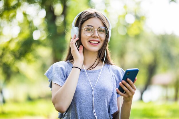 Sonriente joven estudiante con mochila sosteniendo teléfono móvil, caminando en el parque, escuchando música con auriculares