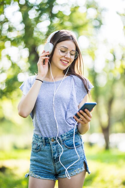 Sonriente joven estudiante con mochila sosteniendo teléfono móvil, caminando en el parque, escuchando música con auriculares