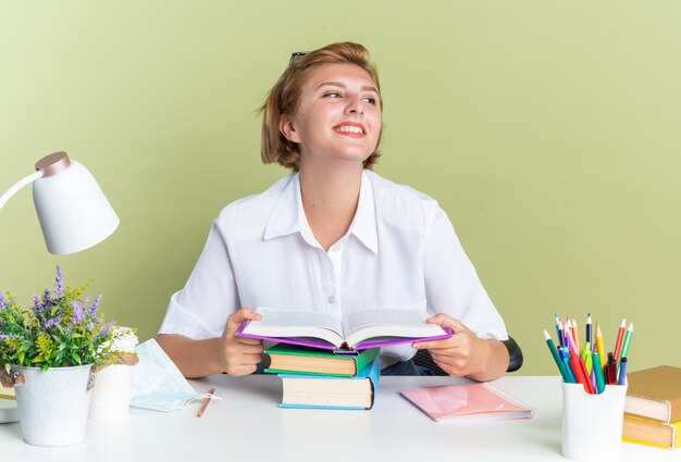 Sonriente joven estudiante rubia sentada en un escritorio con herramientas escolares sosteniendo libro abierto mirando al lado