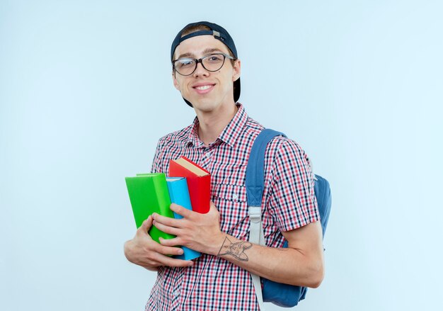 Sonriente joven estudiante vistiendo bolso trasero y gafas y gorra sosteniendo libros en blanco