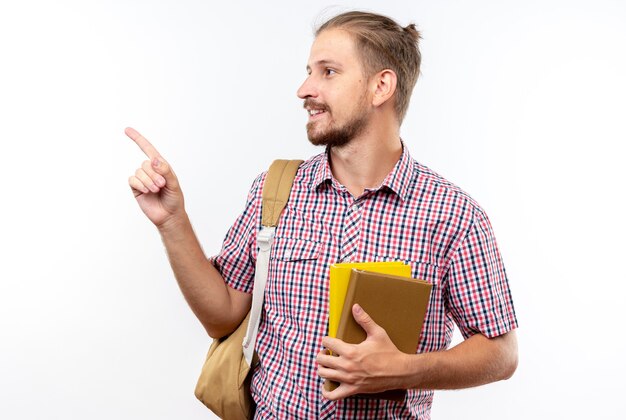 Sonriente joven estudiante vistiendo mochila sosteniendo libros apunta al lado aislado en la pared blanca con espacio de copia