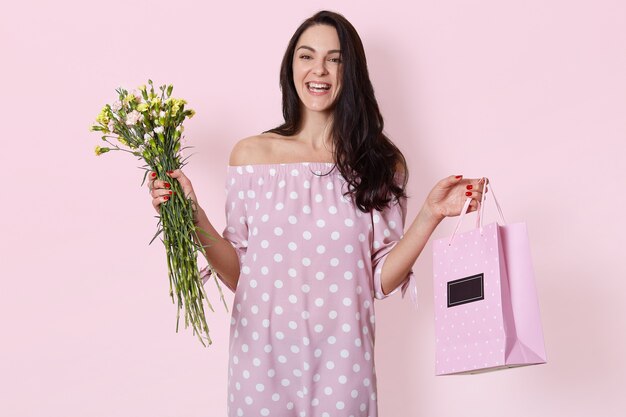 sonriente joven europea tiene el pelo largo y ondulado oscuro, lleva un vestido rosa de lunares, sostiene una bolsa de regalo y un ramo de flores, posa en rosa claro, cumple años.