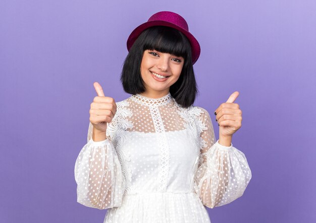 Sonriente joven fiestera con gorro de fiesta mostrando los pulgares para arriba aislado en la pared púrpura