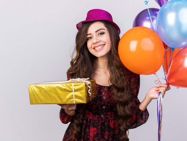 Sonriente joven fiestera con gorro de fiesta sosteniendo globos y paquete de regalo mirando a la cámara aislada en la pared blanca