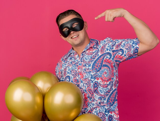 Sonriente joven fiestero con antifaz de mascarada sosteniendo globos y apunta a sí mismo aislado en rosa