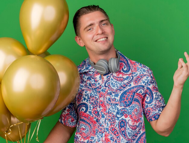 Sonriente joven fiestero con camisa colorida y auriculares alrededor del cuello sosteniendo globos extendiendo la mano aislada en verde