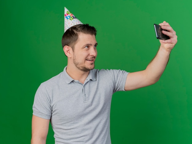 Sonriente joven fiestero con gorro de cumpleaños tomar un selfie aislado en verde