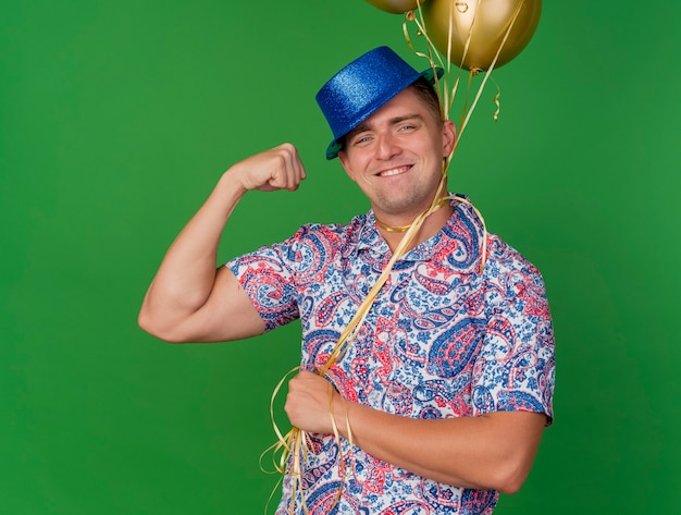 Sonriente joven fiestero con sombrero azul sosteniendo globos atados alrededor del cuello aislado sobre fondo verde