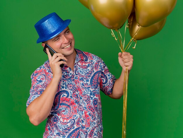 Sonriente joven fiestero con sombrero azul sosteniendo globos y habla por teléfono aislado en verde