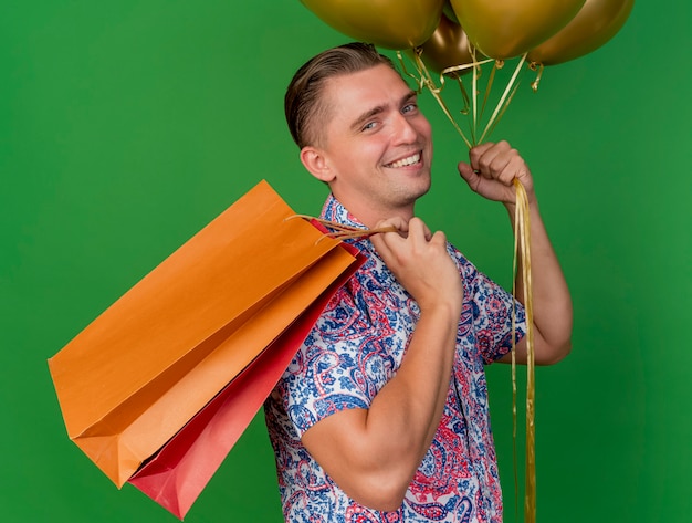 Sonriente joven fiestero con sombrero azul sosteniendo globos poniendo bolsas de regalo en el hombro aislado en verde