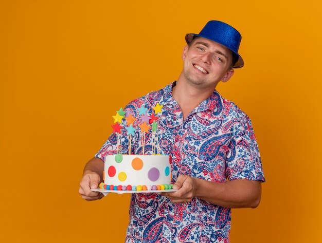 Sonriente joven fiestero con sombrero azul sosteniendo pastel aislado en naranja