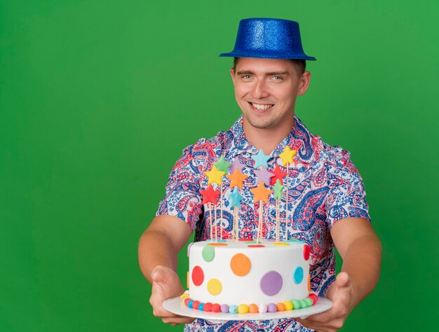 Sonriente joven fiestero con sombrero azul sosteniendo pastel aislado en verde