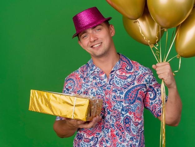 Sonriente joven fiestero con sombrero rosa sosteniendo globos con caja de regalo aislada en verde