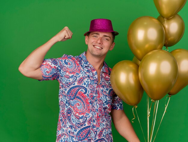 Sonriente joven fiestero con sombrero rosa sosteniendo globos y mostrando un gesto fuerte aislado en verde